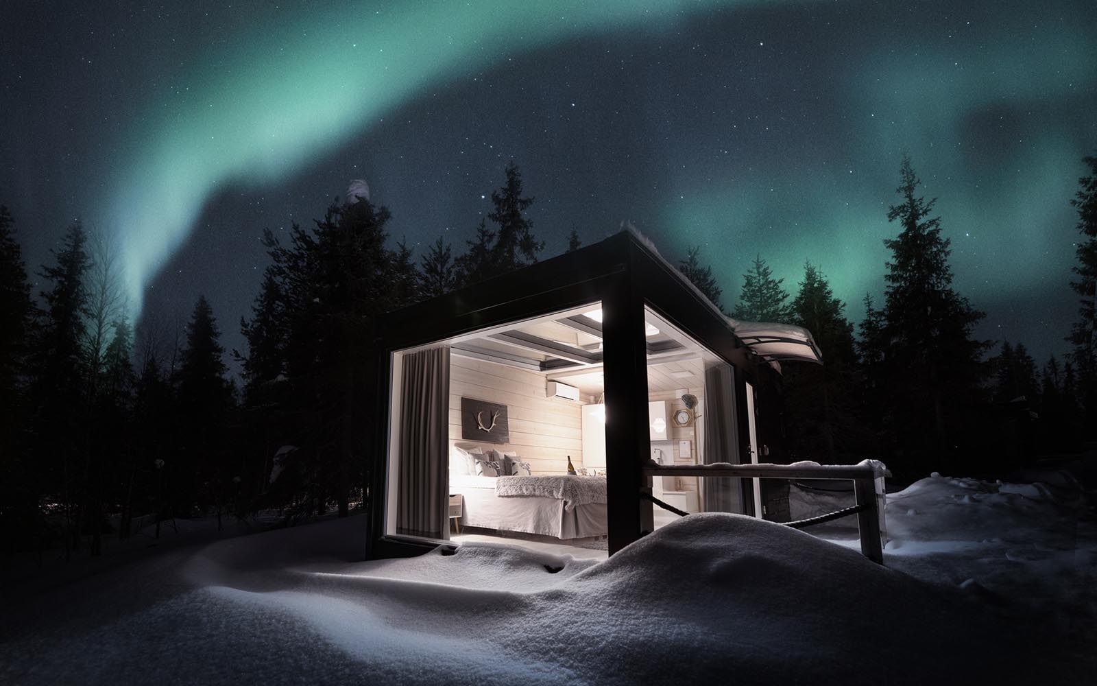 A view across undulating snow towards the lit up interior of the Aurora Glass Villa's at Iisakki Glass Village in Finnish Lapland.  The dark night sky is streaked with turquoise green auroras.