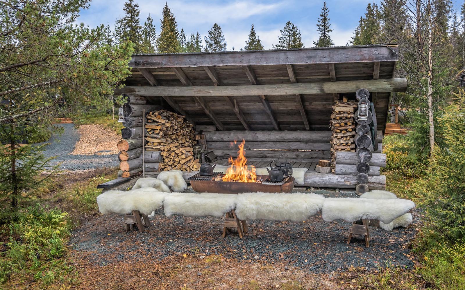 Wooden benches covered with reindeer skins are positioned around the roaring fire pit at an outdoor wooden laavu shelter, stacked with fire wood, at Iisakki Village in Lapland, Finland