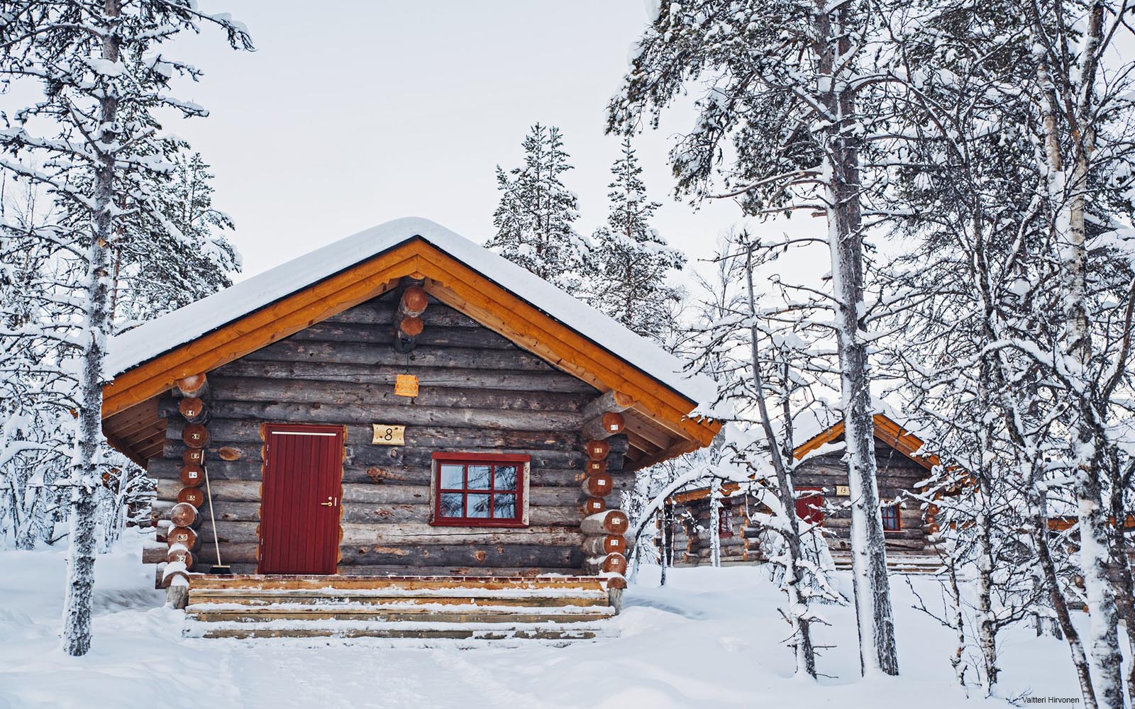 A traditional small log cabin with a red door sits in a snowy setting in East Village at Kakslauttanen Arctic Resort in Finnish Lapland
