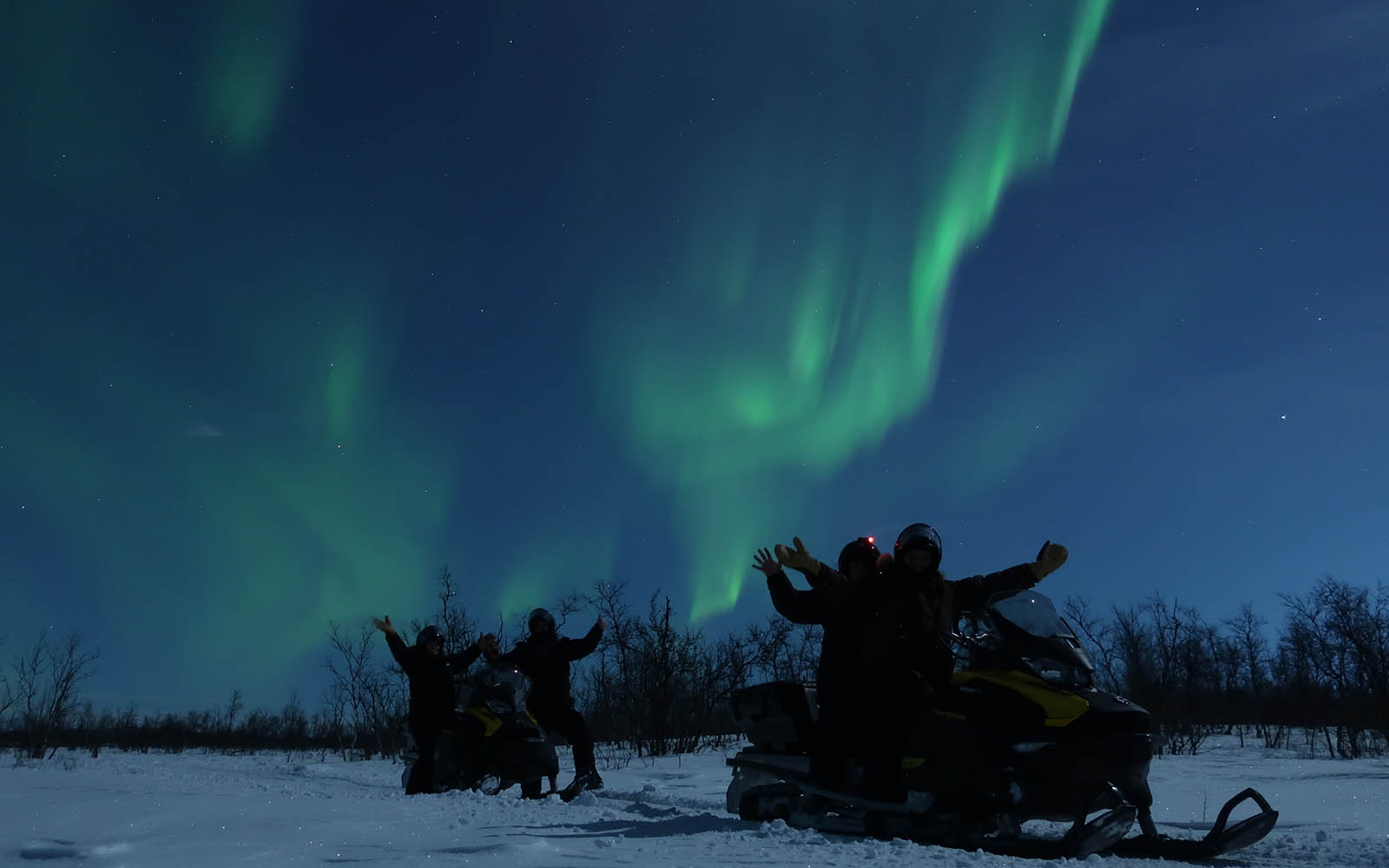 Snowmobile riders wave their hands in the air under a ribbon of green northern lights during a Aurora snowmobile ride in Swedish Lapland 