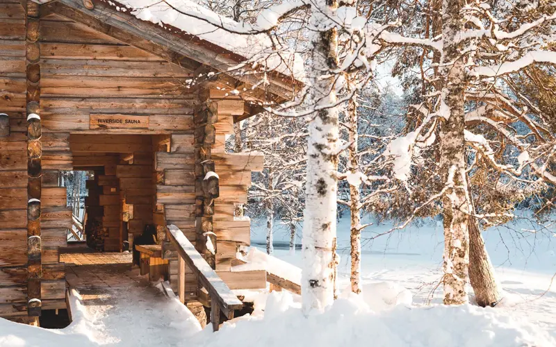 a traditional log sauna building sit in a snowy forest landscape on the edge of the river at Vaattunki Wilderness Resort near Rovaniemi in Finnish Lapland