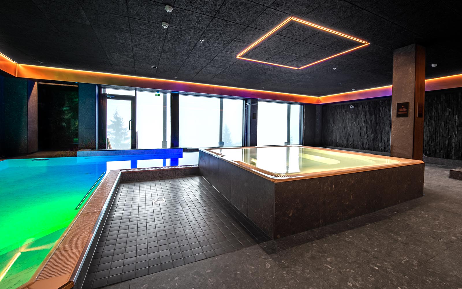 The spa area at Hotel Iso Syote with jacuzzi in the centre.  There are blue and green lights inside the swimming pool, and pink and purple lights edge the dark ceiling mimicing the Northern lights that can often be seen though the large picture window