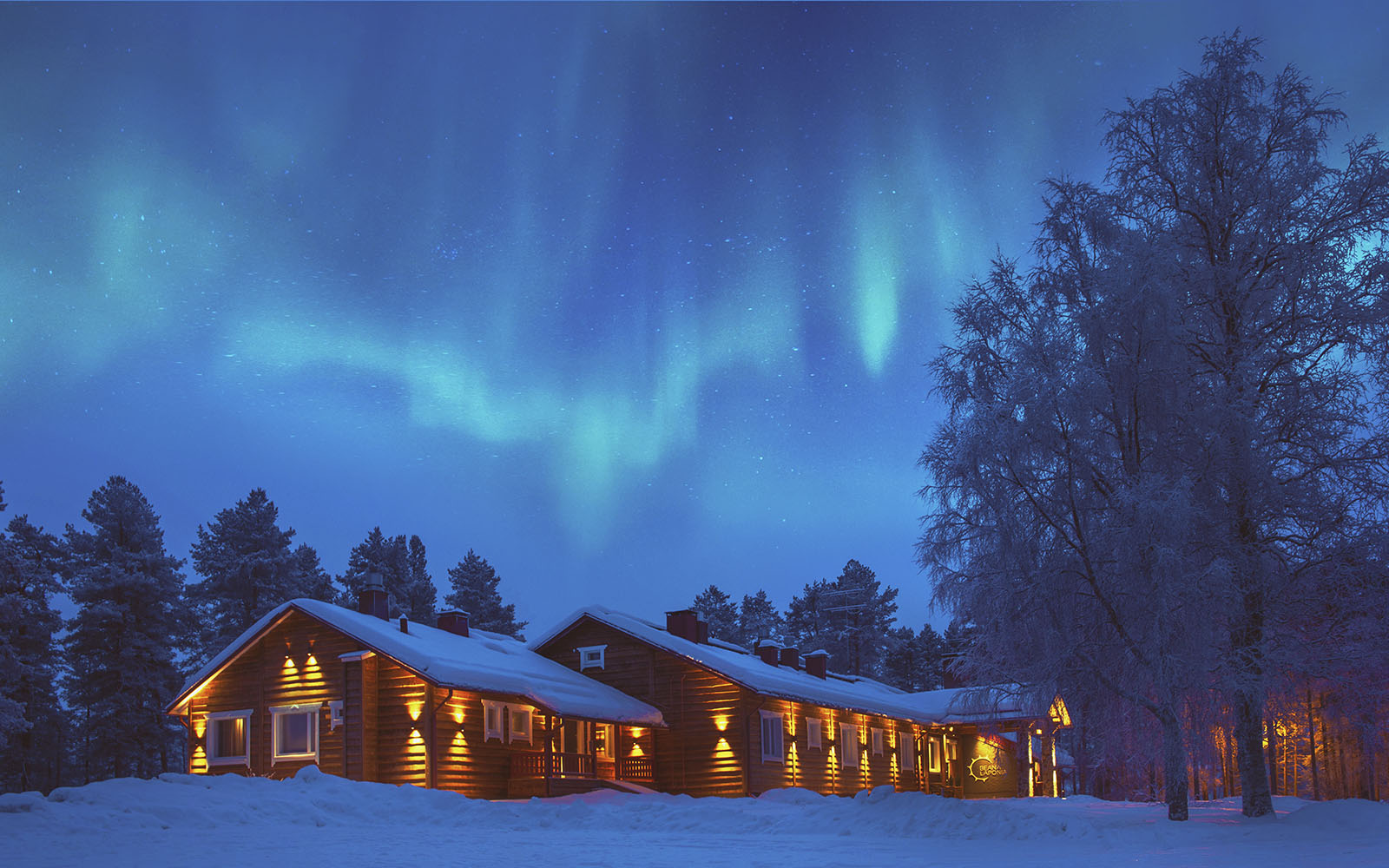 White Northern Lights (aurora borealis) dance across a blue sky above Beana Laponia near Rovaniemi in Finnish Lapland.  The wood clad lodge building is lit with a warm orange glow from the external lights mounted around the walls. 