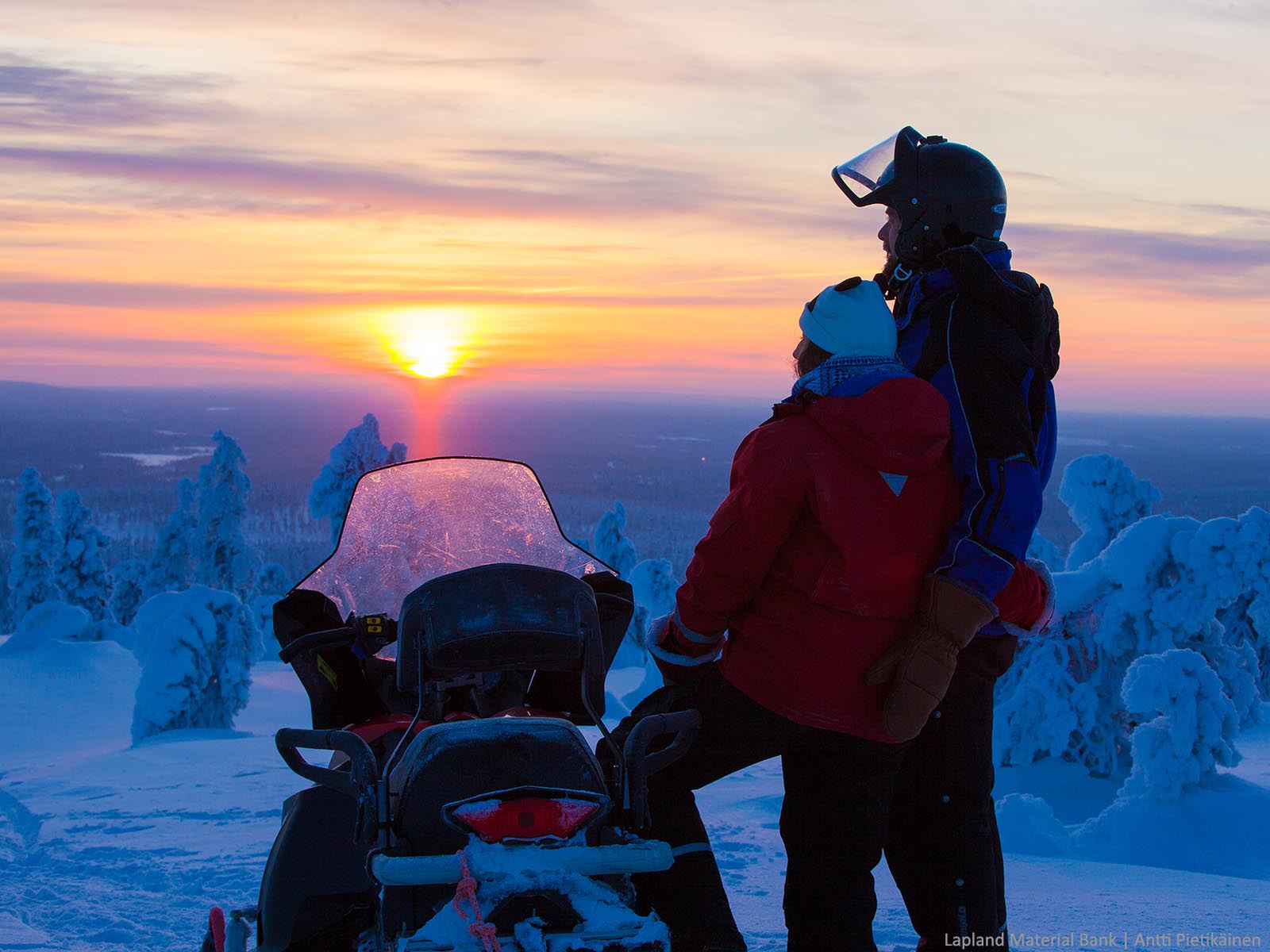 A couple stand holding each other next to a snowmobile and watch the sunset over the snow covered fell in Muonio, Finnish Lapland 