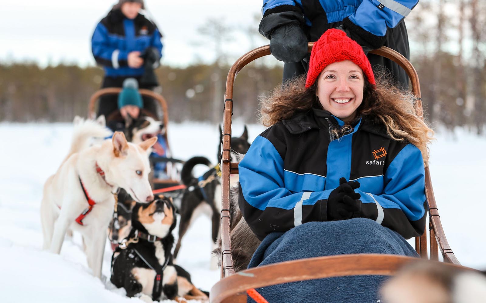 a smiling lady in a red bobble hat riding as a passenger in the sled on a husky safari in Finnish Lapland