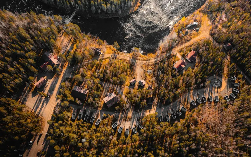 An aerial drone view of Vaattunki Wilderness Resort showing the Panorama Cabins and river rapids in the midnight sun in Lapland.  There are long shadows cast by the tall pine trees that surround the cabins