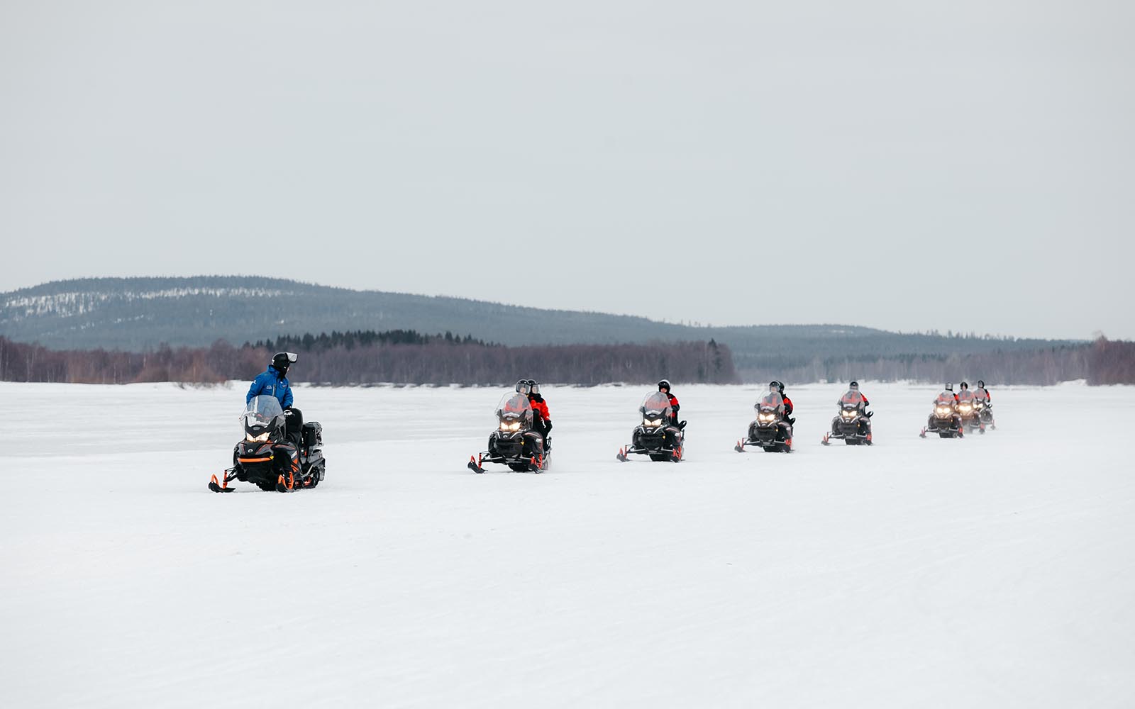 8 snowmobiles ride in a line across a frozen lake lined with tall trees and a fell in the background.  The guide is dressed in blue and the guest all wear red thermal suits
