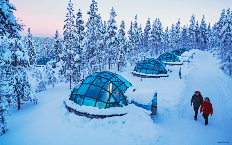 A couple walks along a snowy pathway lined with the famous glass igloos at Kakslauttanen Resort in Finnish Lapland