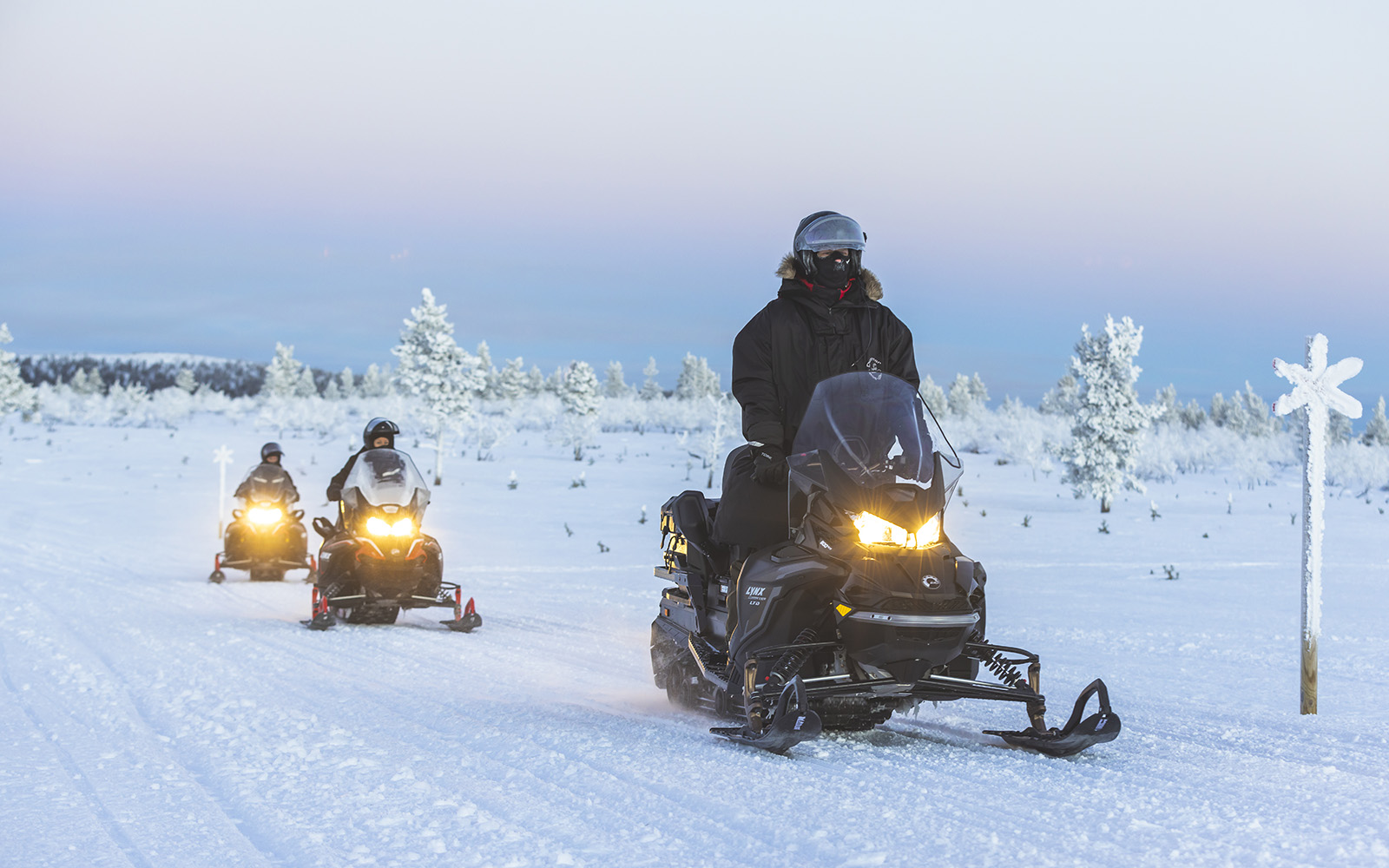 Three black snowmobiles with headlights on are being ridden in a line by solo riders along a snow covered track in Saariselka, Finnish Lapland