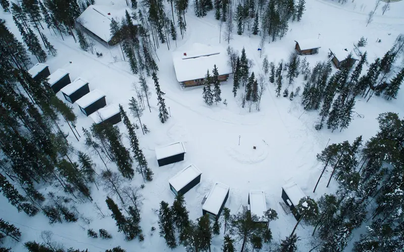 An aerial drone image of the panorama cabins at Vaattunki Wilderness Resort in Finland, surrounded by snow and tall pine trees