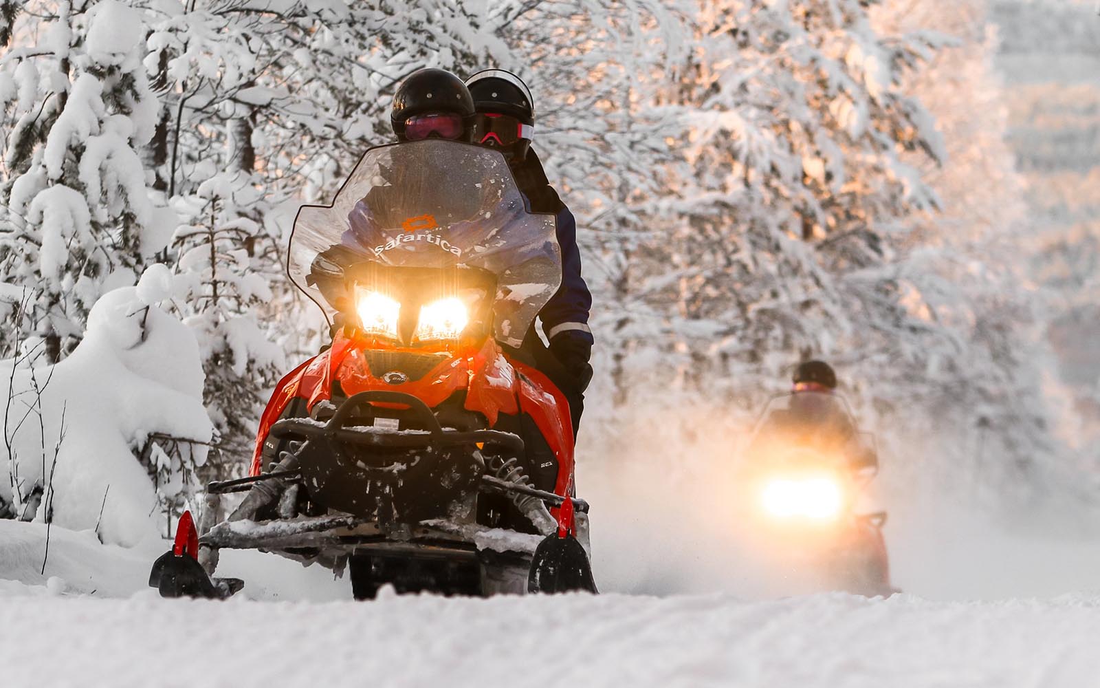two adventurers riding a red snowmobile with headlights on in a snow covered wilderness in Finnish Lapland 