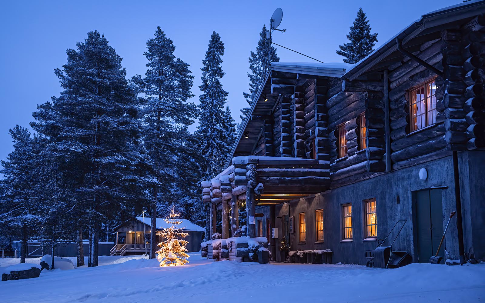 Orange lights glow through the windows of the kelo log constructed main building at Brandon Lodge and a lit Christmas trees sits in the snow in front of the main entrance. 