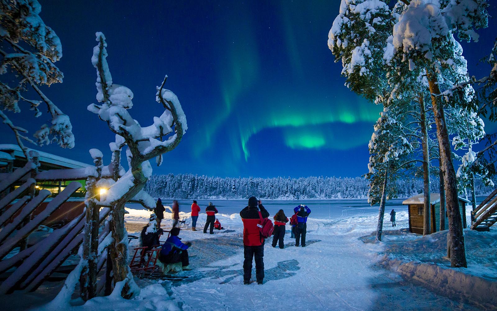Guests stand in the snow outside Harriniva Adventure Resort and take photographs of the northern lights (aurora borealis) as it dances in the bright blue night sky
