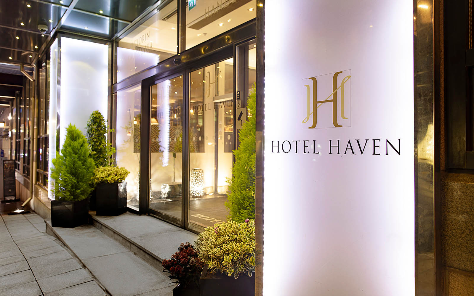 Wide steps, framed by potted shrubs, lead to the glass sliding doors of Hotel Haven Helsinki, the hotel logo features on the frosted glass façade