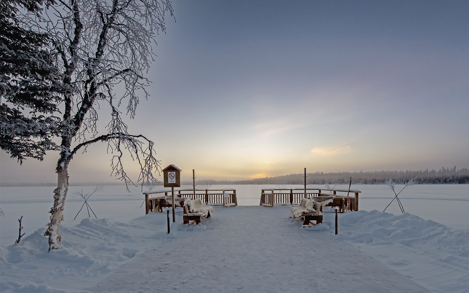 Wooden benches draped with reindeer skins sit on a snow covered deck that leads to a frozen lake and ice hole at Nivunki Village in Finnish Lapland 