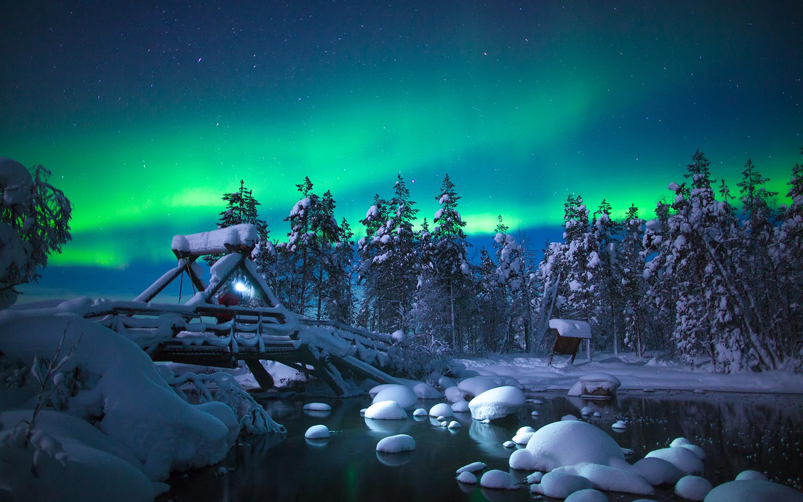 The Northern Lights (aurora borealis) streaks across the blue night sky over a snow covered bridge, snow covered rocks sit in the water