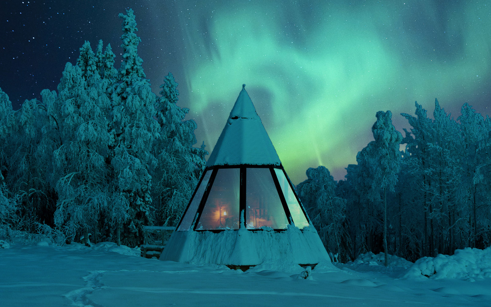 A pyramid shaped Aurora Kota Hut, covered in snow, sits in the blue light of polar night at Nivunki Village in Finnish Lapland.  The Northern Lights swirl in the sky above the hut while the interior lights emit a warm glow from inside