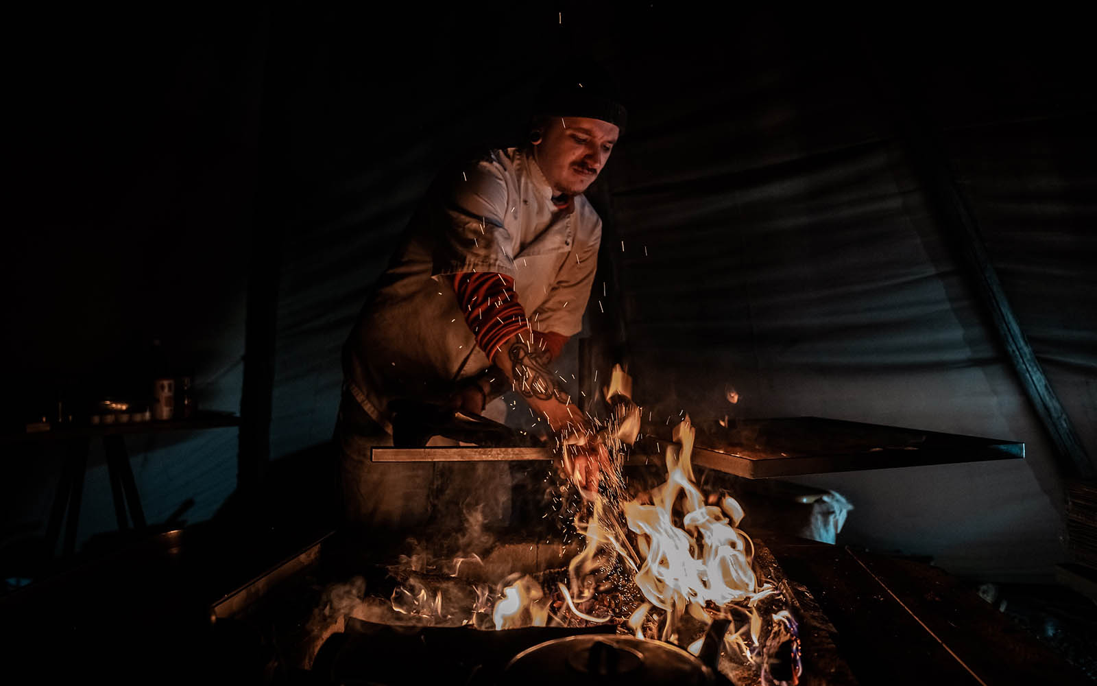 A tattooed chef prepares a BBQ dinner over a roaring open fire in the tipi at Mattarahkka Northern Lights Lodge near Kiruna in Northern Sweden