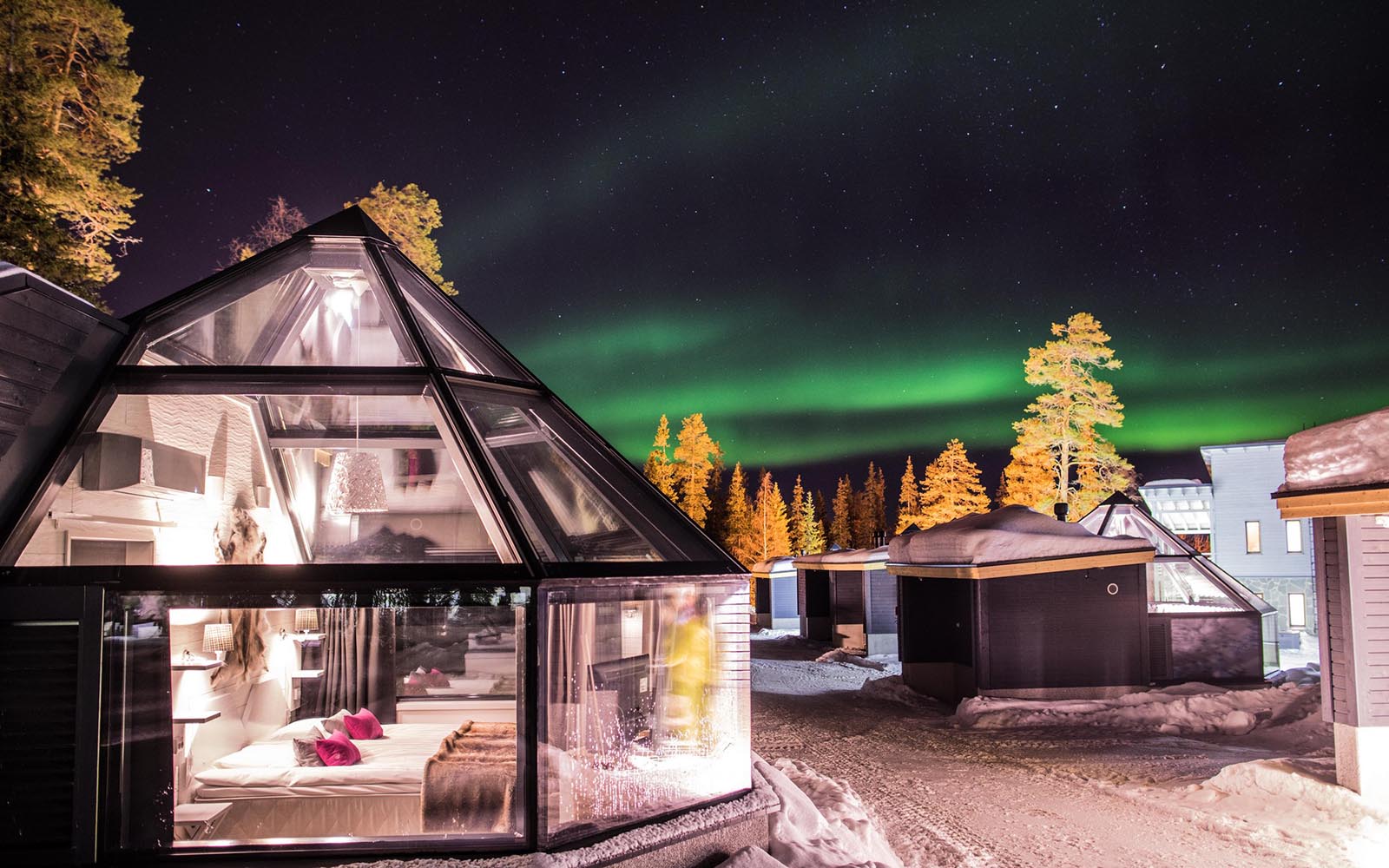 Premium glass igloos with a pyramid shaped glass roof at Santa's Hotel Aurora in Luosto, Finnish Lapland sit in snowy grounds under the northern lights 