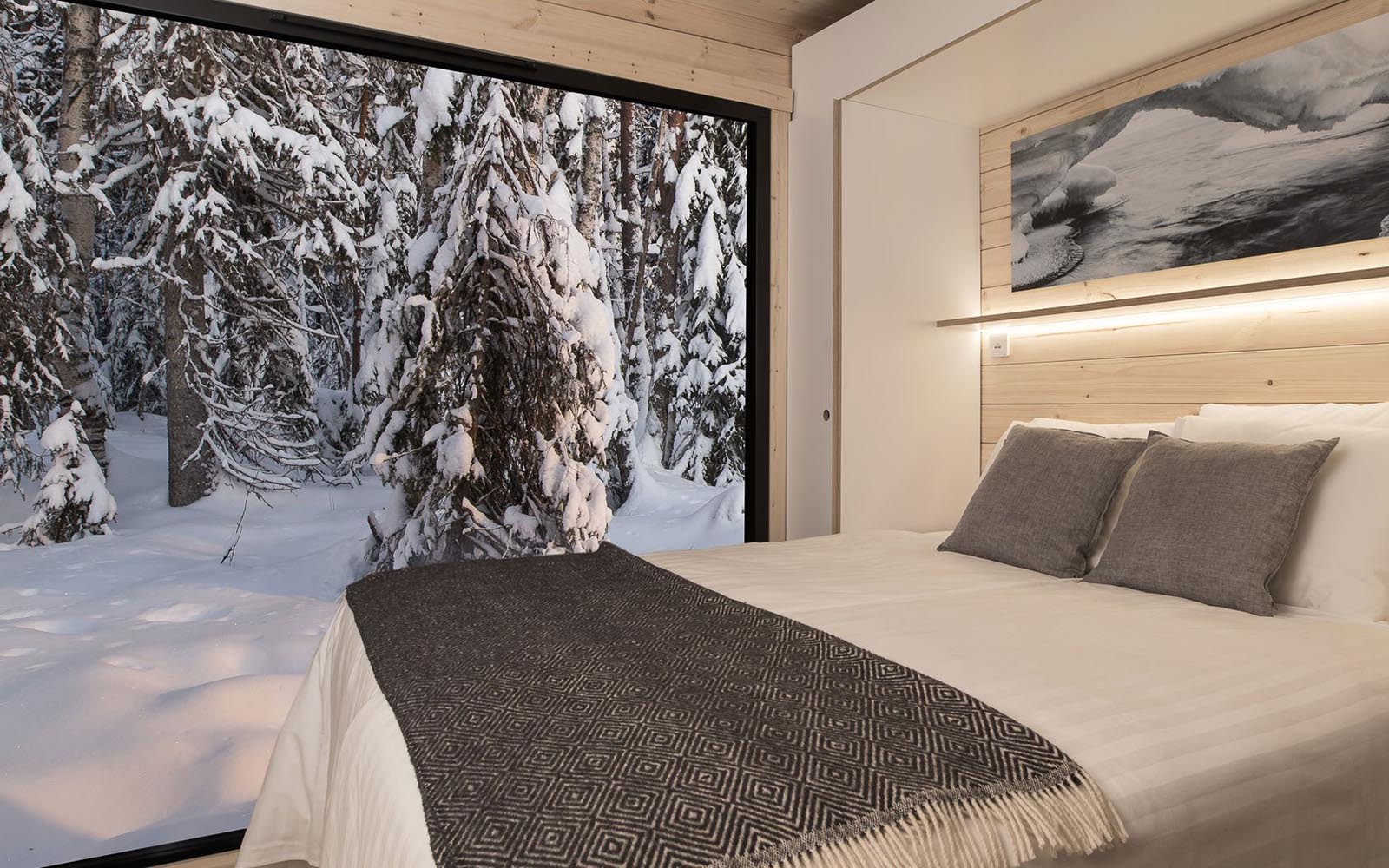 A double bed with crisp white linens, grey cushions and a grey woollen throw, next to a panoramic floor to ceiling window in the panorama cabin at Vaattunki Wilderness Resort