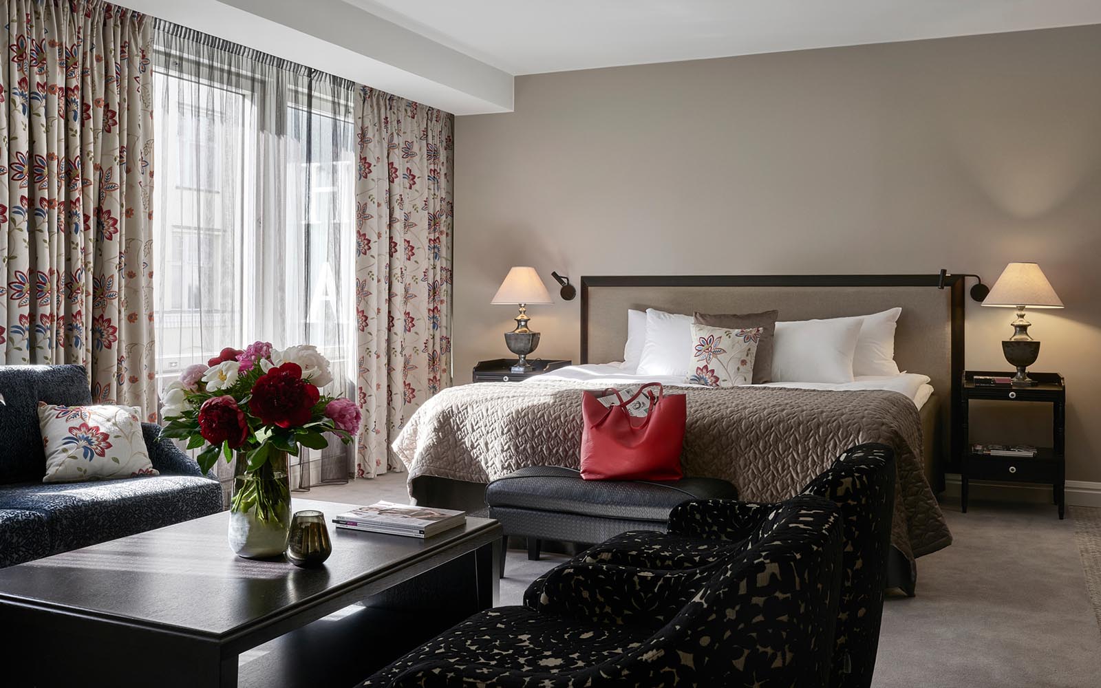 A tastefully decorated guest suite at Hotel Haven, Helsinki with floral curtains, a king bed with twin nightstands and a seating area with comfortable sofa, two armchairs and a coffee table 