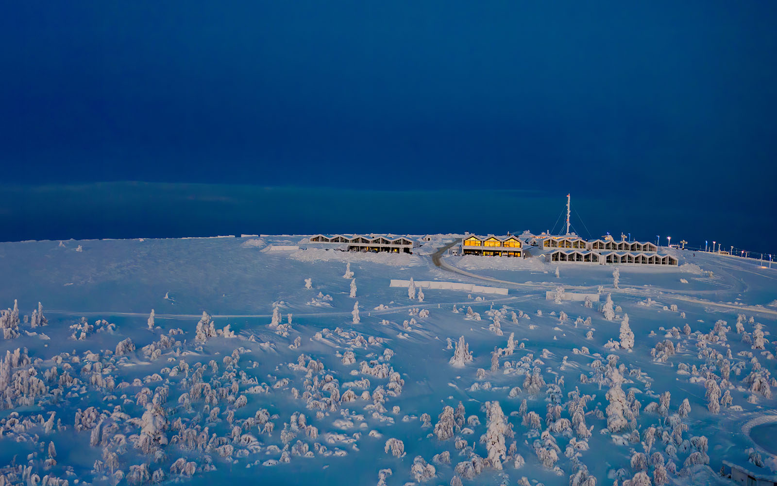 A distant shot of Star Arctic Resort nested on top of the snow covered fell in Saariselka. The resort buildings are lit with a warm orange glow which contrasts with the blue light of the polar night in Lapland 
