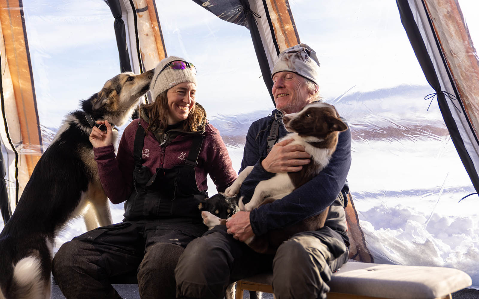 A man cuddles a husky and another dog commands attention from the lady in a tented lavvu at Husky Home in Kiruna, Swedish Lapland