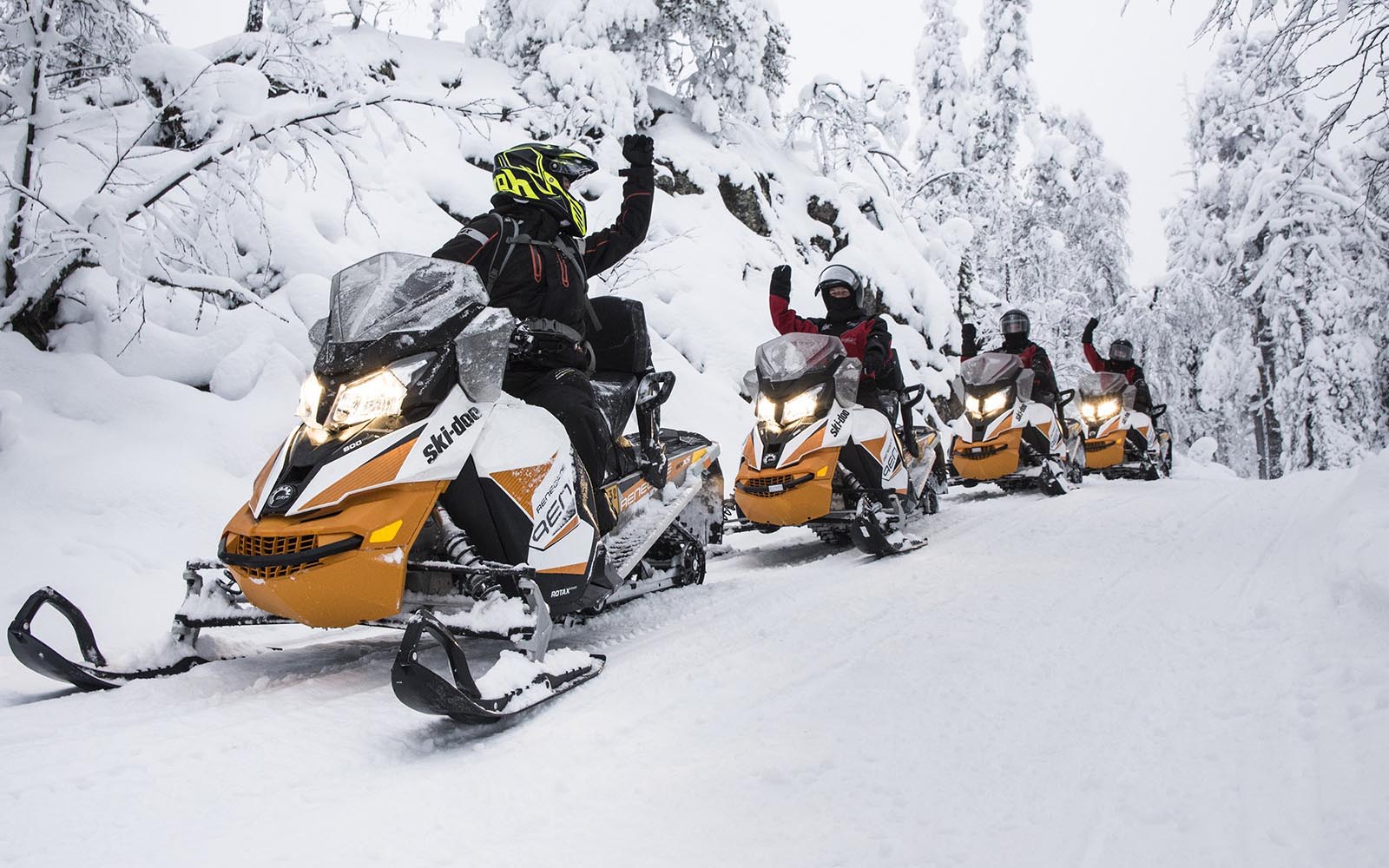4 riders on yellow and white snowmobiles in a snowy setting indicate to the lead rider that they are all ready to go by raising their arm.