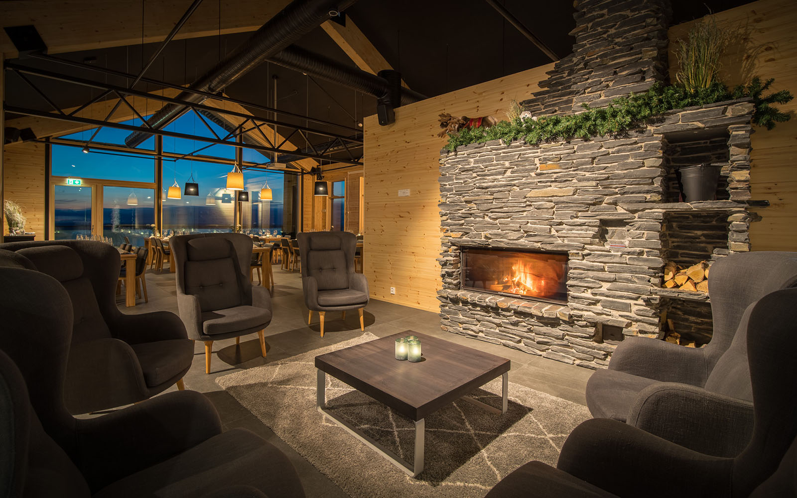 An artist's impression the new lounge area at Star Arctic Resort in Saariselka, Finland. Comfortable high backed arm chairs sit under a vaulted wooden ceiling around a coffee table in front of a large stone fireplace and far reaching views can be seen in the background though the huge panoramic windows